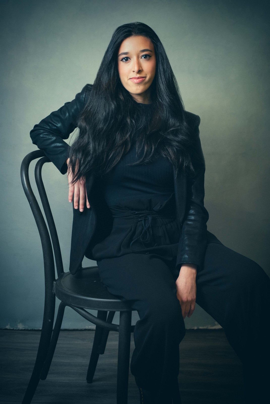 A woman with a medium-dark skin with long dark hair, wearing a black long-sleeve shirt and black pants seated in a black chair, one arm is resting on the back of the chair and the other is draped into her lap, in a gray room with a grayish wood floor.